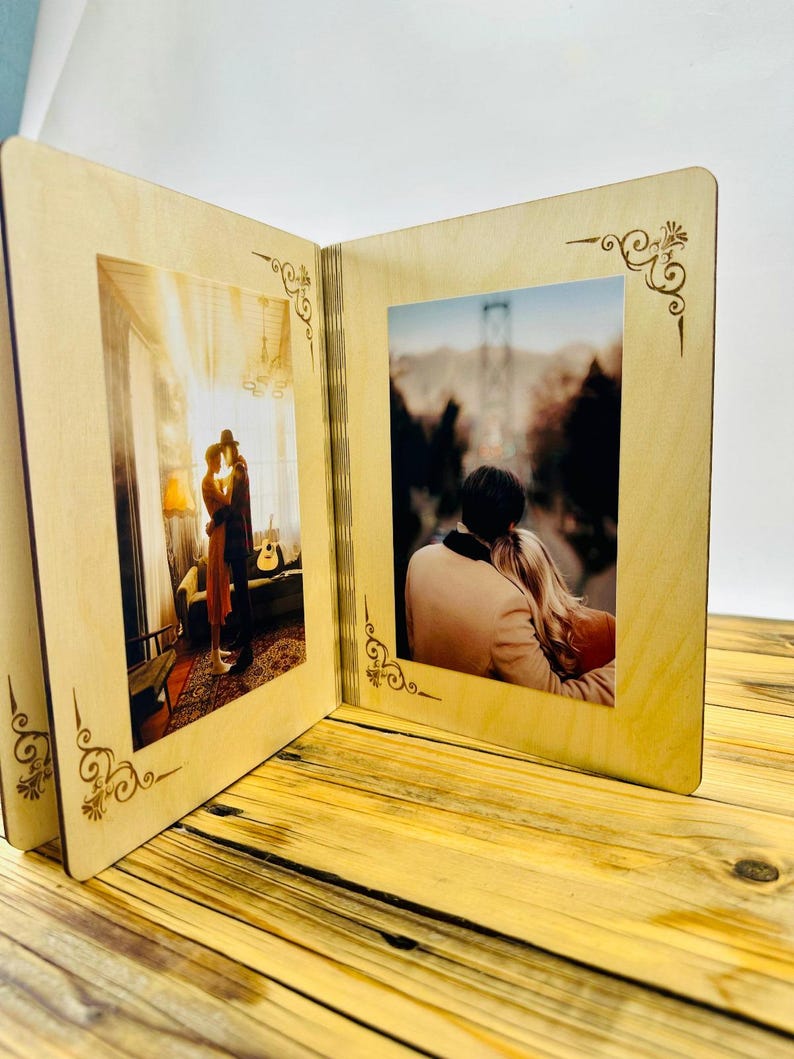 May include: A wooden photo album with two open pages, each displaying a photograph of a couple. The album has a light brown wood finish and intricate decorative designs around the edges of the photo frames.