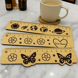 Puede incluir: Tres reglas de madera con diferentes diseños. La regla superior tiene un sol, una estrella fugaz y planetas. La regla del medio tiene flores y el texto "Your Name". La regla inferior tiene mariposas y flores.