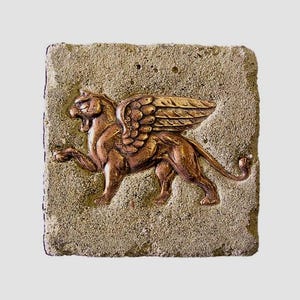 Puede incluir: Escultura en relieve de león alado de color bronce sobre una piedra cuadrada texturizada de color marrón claro. El león ruge, con alas detalladas y una cola enroscada. La piedra tiene un aspecto rugoso y envejecido.