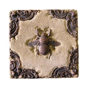 May include: Square, beige tile featuring a raised, bronze-coloured bee in the centre. The tile has a decorative bronze border with ornate scrollwork in each corner. The bee has detailed wings and body segments.