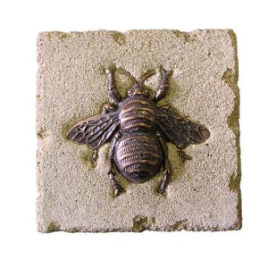Puede incluir: Una escultura de abeja de color bronce está fijada a una piedra cuadrada, texturizada y de color beige claro. La abeja tiene alas, patas y antenas detalladas. La piedra tiene un borde ligeramente irregular, lo que le da un aspecto rústico.