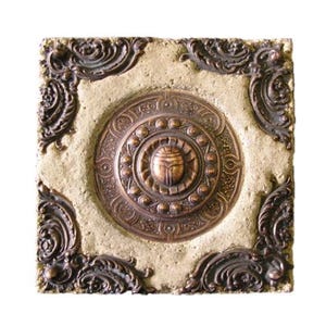 Könnte beinhalten: Quadratische, strukturierte Fliese mit einem zentralen, bronzefarbenen Medaillon mit einem ägyptischen Skarabäus-Käfer-Design. Das Medaillon ist von einem kreisförmigen Muster umgeben und von verzierten Eckakzenten in passendem Bronzeton umrahmt. Der Hintergrund ist hellbeige.