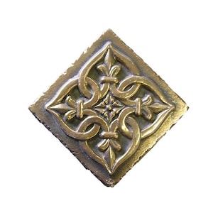 Puede incluir: Una baldosa decorativa cuadrada de color bronce con un diseño ornamental en relieve. El motivo central es una forma de trébol de cuatro hojas con elementos de flor de lis y bucles entrelazados. La baldosa tiene una superficie texturizada y un aspecto ligeramente envejecido.