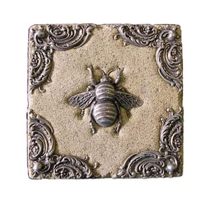 May include: A square, beige tile with a raised, detailed bee in the center. The tile is framed by ornate, silver-colored corner accents. The bee has striped detailing on its body and wings.