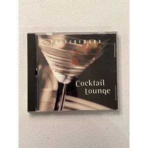 May include: A CD case featuring the Pottery Barn logo and the words "Cocktail Lounge." The cover art displays a martini glass with olives and a cherry. The case is black with a clear plastic cover.