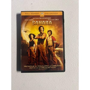Peut inclure: Boîtier DVD du film "Sahara" avec Matthew McConaughey, Steve Zahn et Penélope Cruz. La couverture présente un thème désertique avec le titre en grandes lettres dorées. Le boîtier DVD est noir avec les mots "Full Screen Collection" en haut.