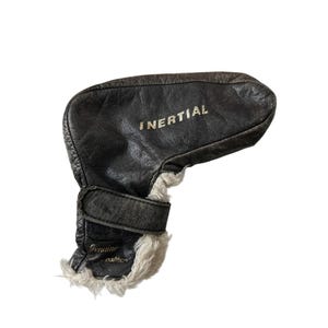 Könnte beinhalten: Eine schwarze Golfschlägerhaube aus Leder mit dem Wort "INERTIAL" auf der Oberseite. Die Haube hat ein weißes, flauschiges Innenfutter und einen Riemenverschluss. Die Worte "Genuine leather" sind auf der Seite aufgedruckt.