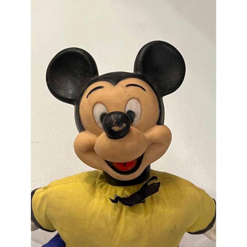 Op de afbeelding: Vintage Mickey Mouse pop met zwarte oren, een zwarte neus en een lachend gezicht. De pop draagt een geel shirt met zwarte bies en een zwarte vlinderdas. De armen van de pop zijn wit.