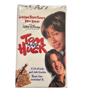May include: A Tom and Huck VHS tape featuring Jonathan Taylor Thomas and Brad Renfro on the cover. The title is printed in large red letters with Walt Disney Pictures Presents above it. A map with a knife and the text A lot of kids get into trouble. These two invented it. are visible.