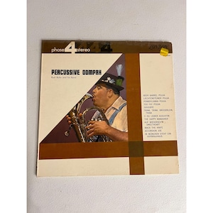 May include: A vintage vinyl record album titled "Percussive Dompah" by Rudi Bohn and his Band. The album cover features a man playing a brass instrument. The album has a brown and white color scheme with a list of songs on the right side.