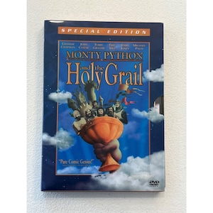 Może przedstawiać: Pudełko DVD do filmu "Monty Python and the Holy Grail" z niebieską okładką, na której widnieje tytuł filmu i nazwiska aktorów. Grafika zawiera złoty kielich, zamek i postacie w średniowiecznych strojach. Widoczny jest również tekst "Pure Comic Genius!".