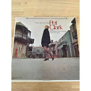 Peut inclure: Pochette d'album vinyle vintage pour "These Are My Songs" de Pet Clark. La couverture présente une femme en tenue noire marchant dans une scène de rue. Le titre de l'album et le nom de l'artiste sont imprimés en rouge et blanc.