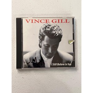 Könnte beinhalten: Eine CD-Hülle mit Vince Gill und dem Albumtitel "I Still Believe In You". Das Cover ist ein Schwarz-Weiß-Foto von Vince Gill. Die CD-Hülle ist signiert. Der Text "VINCE GILL" ist rot.