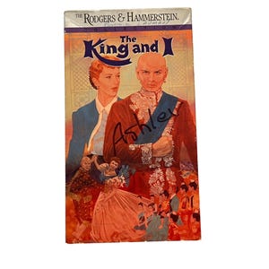 May include: A VHS tape cover for The Rodgers & Hammerstein Collection, The King and I. The cover features an illustration of a man and woman, with the name Ashley written in black marker across the center and handwritten text at the top.