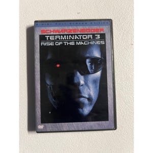 May include: A DVD case for "Terminator 3: Rise of the Machines" featuring Arnold Schwarzenegger. The cover art shows a close-up of a cyborg's face with a red eye and the film's title in red and white text. The DVD case is black.
