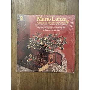 May include: A vintage vinyl record album titled "Christmas Hymns and Carols" by Mario Lanza. The album cover features a festive arrangement of holly and berries, a framed portrait, and a book. The album is sealed in plastic.