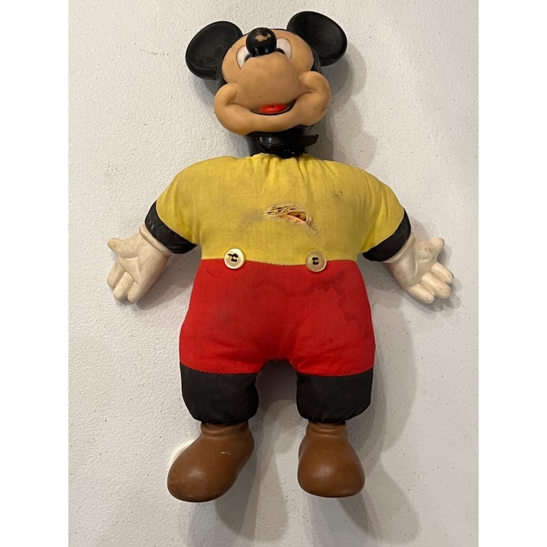 Op de afbeelding: Vintage Mickey Mouse pop met een rubberen gezicht en zwarte oren. De pop draagt een geel shirt met zwarte mouwen, een rode korte broek en bruine schoenen. De shirt heeft twee knopen en een scheur. De handen zijn wit.