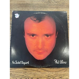 以下が含まれることがあります： 赤みがかったオレンジ色の男性の顔のクローズアップが特徴のビニールレコードアルバムのカバー。「No Jacket Required」というアルバムタイトルとアーティスト名「Phil Collins」が白で印刷されています。