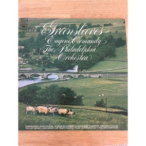 以下が含まれることがあります： ユージン・オーマンディとフィラデルフィア管弦楽団による「Greensleeves」のヴィンテージビニールレコードアルバムのカバー。カバーには、橋、羊、緑の丘がある風景が描かれています。アルバムのトラックリストは下部に記載されています。