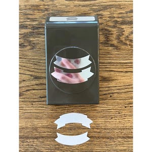 May include: A dark gray banner-shaped paper punch with a clear window showing the banner design. A cut-out white banner is below the punch on a wooden surface. The punch is designed for crafting and scrapbooking.