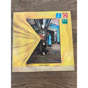 May include: A vintage vinyl record album cover featuring a yellow curtain partially pulled back to reveal a band of four people standing in a colorful, futuristic hallway. The album title "SHEET MUSIC" is printed at the bottom of the cover. The album cover also features the band's name "10 CC" in a colorful block design.