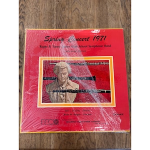 Pode incluir: Uma capa de álbum de vinil vermelho com uma foto de um homem tocando oboé. O álbum é intitulado "Spring Concert 1971" e apresenta a Roger B. Taney High School Symphonic Band. O álbum é em estéreo.