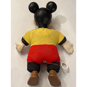 Op de afbeelding: Vintage Mickey Mouse trekspeelgoed. Het speelgoed heeft een zwarte kop met grote oren, een geel shirt, een rode broek en bruine schoenen. De armen en handen zijn wit. Aan de achterkant is een trekkoord bevestigd.
