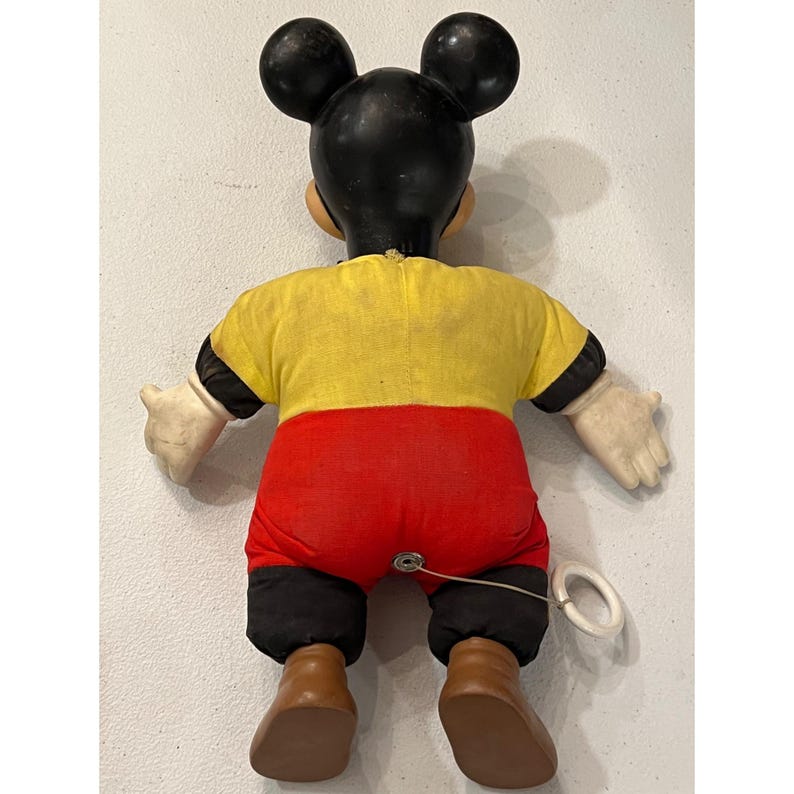 Op de afbeelding: Vintage Mickey Mouse trekspeelgoed. Het speelgoed heeft een zwarte kop met grote oren, een geel shirt, een rode broek en bruine schoenen. De armen en handen zijn wit. Aan de achterkant is een trekkoord bevestigd.