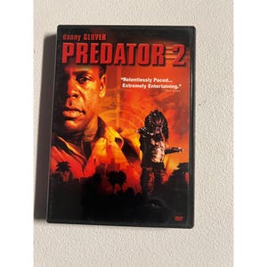 May include: DVD case for the movie "Predator 2" featuring Danny Glover. The cover art displays Glover's face and a Predator figure against a red and orange background. Text includes the movie title and a quote.