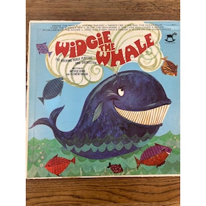 May include: Vintage vinyl record album cover featuring a smiling blue whale with white teeth, surrounded by colorful fish. The title "Widgie the Whale" is in large red letters. The album cover has a light blue background with the text "The Rocking Horse Players and Orchestra."