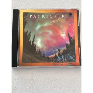 May include: A CD case featuring the album "Sapphire" by Patrick Ki. The cover art shows a vibrant aurora borealis scene with a forest silhouette. The artist's name and album title are prominently displayed.