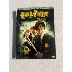 Puede incluir: DVD de "Harry Potter y la Cámara de los Secretos" con el texto "Full-Screen Edition". La portada presenta a Harry Potter, Ron Weasley y Hermione Granger, con Dobby y una espada. La caja del DVD es negra con la parte superior verde.