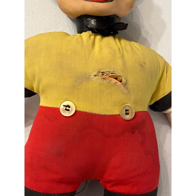 Op de afbeelding: Vintage stoffen pop met een geschilderd gezicht, zwarte kraag en een geel shirt met een gat. De pop heeft een rode broek en zwarte schoenen. Twee gouden knopen bevestigen het shirt. Het gezicht van de pop is beschilderd met rode lippen en zwarte ogen.