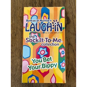 May include: A rectangular DVD case for Rowan & Martin's Laugh-In The Sock-It-To-Me collection. The cover features a vibrant yellow, orange, and pink geometric pattern with stylized flower shapes and the text You Bet Your Bippy in green.