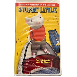 Puede incluir: Cinta de vídeo VHS de la película "Stuart Little". La portada presenta un ratón de dibujos animados con un suéter rojo y zapatillas, de pie sobre un zapato rojo. El título está en azul y amarillo, con el texto "The Little Family Just Got Bigger."