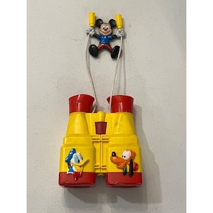 Könnte beinhalten: Ein Vintage-Spielzeugfernglas-Set mit Micky Maus, Donald Duck und Pluto. Das Fernglas ist gelb mit roten Akzenten und Charakterillustrationen. Micky Maus wird dargestellt, wie er das Fernglas von oben hält. Ein klassisches Disney-Sammlerstück.
