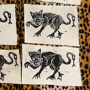 Puede incluir: Seis parches de tela con un diseño de gato negro. El gato está representado en un estilo linograbado estilizado, con una cola larga y una expresión amenazante. Los parches son rectangulares y de tela clara.