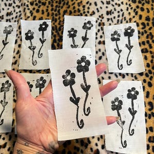May include: A collection of small, rectangular fabric pieces, each featuring a black floral design. The flowers have a simple, stylized appearance. The pieces are arranged on a leopard print background.