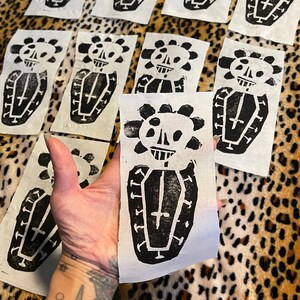 May include: A collection of black and white linocut prints featuring a skull with a floral crown above a coffin. The prints are on rectangular pieces of white fabric. The background is a leopard print.