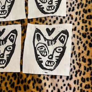Könnte beinhalten: Vier weiße Stoffquadrate mit schwarzen Katzengesicht-Linolschnittdrucken. Die Katzenköpfe haben ein Herz über dem Kopf und eine stilisierte, folkige Ästhetik. Der Hintergrund ist ein Leopardenmuster.