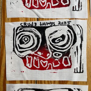 May include: Close-up of three white fabric squares with a black and red abstract face design. The design includes spiral eyes, a red mouth, and the text "CRUDE THINGS 2025". The fabric has a raw, unfinished edge.