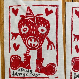 May include: Red linocut print on white fabric featuring a strawberry-like character with a hat, boots, and arms. The print includes the text "CRUDE THINGS '23" and heart shapes. The artwork is displayed on a wooden surface.