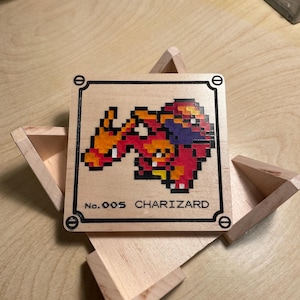 May include: Wooden square featuring a pixelated Charizard design in red, orange, and purple. The artwork is framed with a black border and the text "No. 005 CHARIZARD". The square is set in an open wooden box.