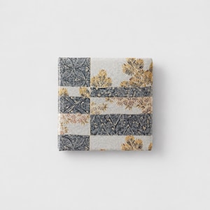 May include: A square decorative box with a patchwork design. The box features alternating sections of patterned fabric in shades of grey, cream, and gold. The patterns include floral and geometric motifs.