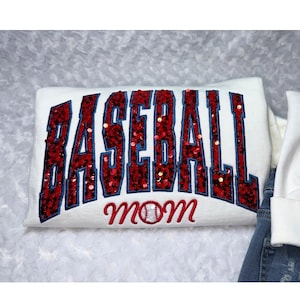 May include: A white sweatshirt featuring the word BASEBALL in large, red sequined block letters with a dark blue outline. Below the word, the word mom is embroidered in red script, with a white baseball graphic replacing the letter o.