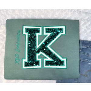 May include: A sage green sweatshirt with a large, sequined letter "K" in emerald green and white. The text "KD Embroidery" is embroidered in green on the left side. The sweatshirt is paired with blue jeans.