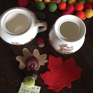 Puede incluir: Dos azucareros de cer&aacute;mica blanca con dise&ntilde;os florales marrones, una hoja de arce de fieltro rojo, una hoja de bellota de fieltro marr&oacute;n, una bellota de fieltro verde y una nota adhesiva amarilla con el texto "1/2 cups x sugar boat - honey pot."
