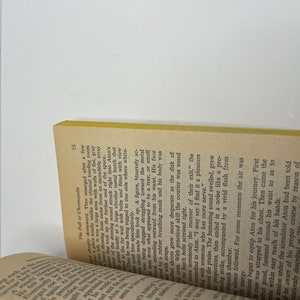 May include: A close-up of a book with a yellow spine and a brown cover. The text on the page is about a character named Aton, who is being prepared for a journey. The text describes the process of equipping Aton for the journey, including strapping a case to his chest, fastening a control panel around his waist, and administering a hypodermic needle.