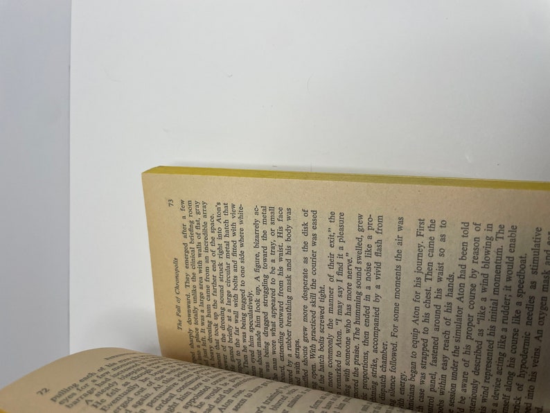 May include: A close-up of a book with a yellow spine and a brown cover. The text on the page is about a character named Aton, who is being prepared for a journey. The text describes the process of equipping Aton for the journey, including strapping a case to his chest, fastening a control panel around his waist, and administering a hypodermic needle.