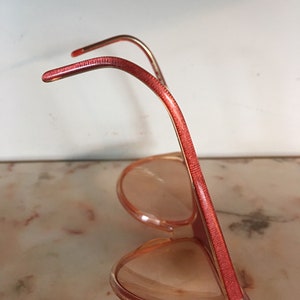 May include: A pair of pink plastic sunglasses with red frames. The sunglasses are in a side view, with the arms of the glasses folded up.